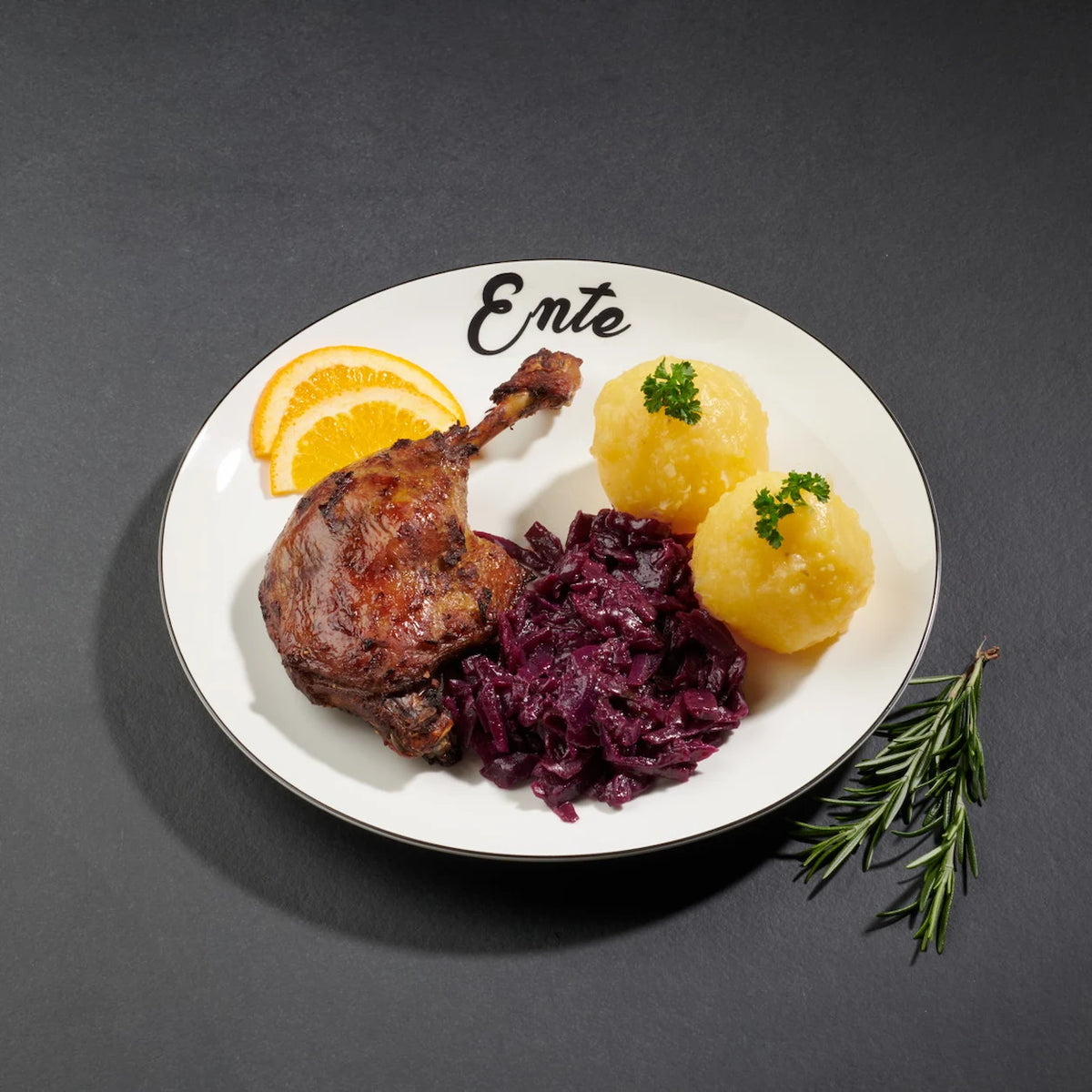 Eine gegarte Entenkeule angerichtet auf einem weißen Teller mit Klößen und Rotkohl und zwei Orangenscheiben als Deko.