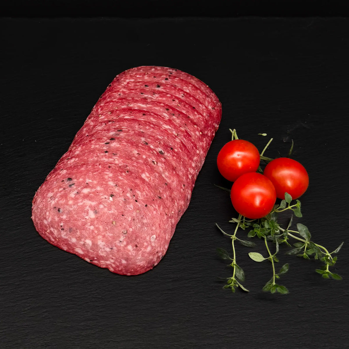 Geschnittene Trüffelsalami, gefächert, auf einer schwarzen Schieferplatte mit Dekoration aus Tomaten und Kräutern.