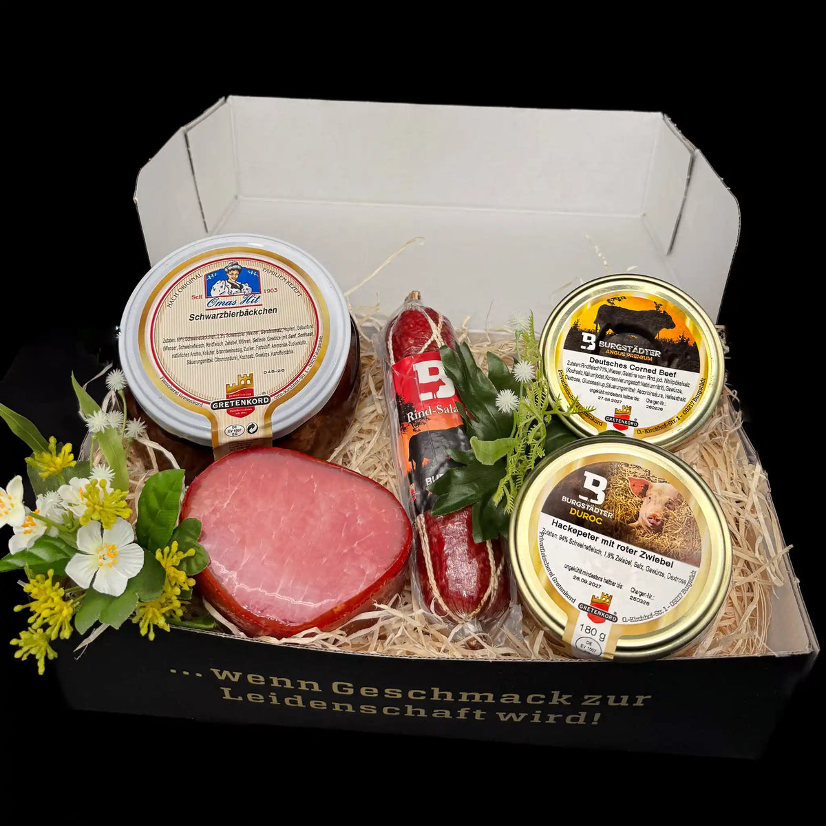 Eine rechteckige Box gefüllt mit einem Glas Corned Beef und einem Glas Hackepeter, einer Salami, einem Lachsschinken und einem Glas Schwarzbierbäckchen.