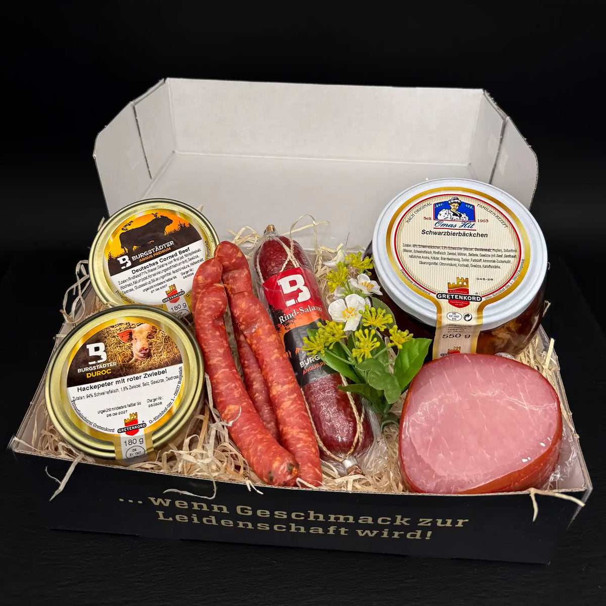 Eine rechteckige Box gefüllt mit einem Glas Corned Beef und einem Glas Hackepeter, einer Salami, 2 Paar Pfefferbeißern, einem Lachsschinken und einem Glas Schwarzbierbäckchen.