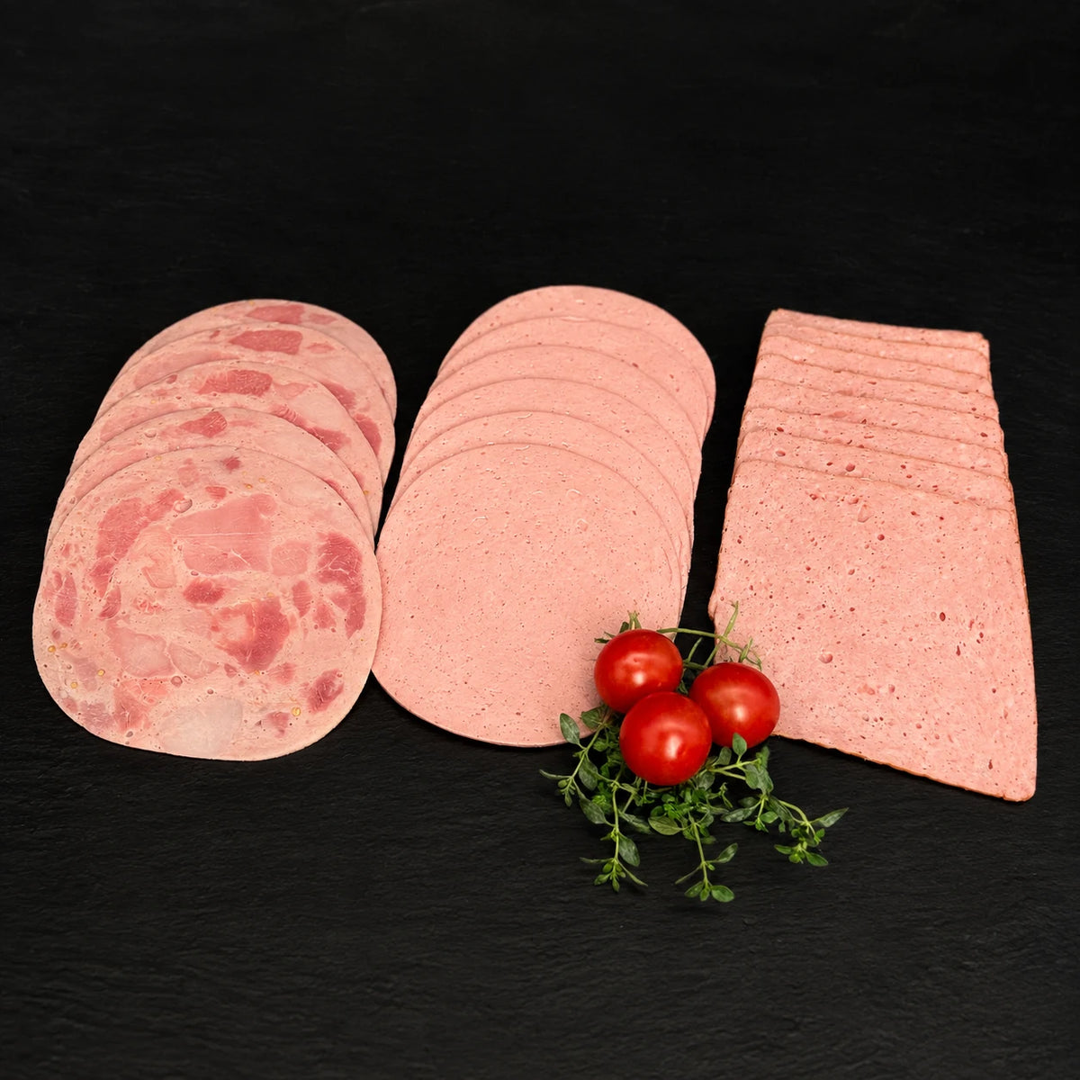 Geschnittener Bierschinken, Leberkäse und Römerbraten gefächert, auf einer schwarzen Schieferplatte mit Dekoration aus Tomaten und Kräutern.