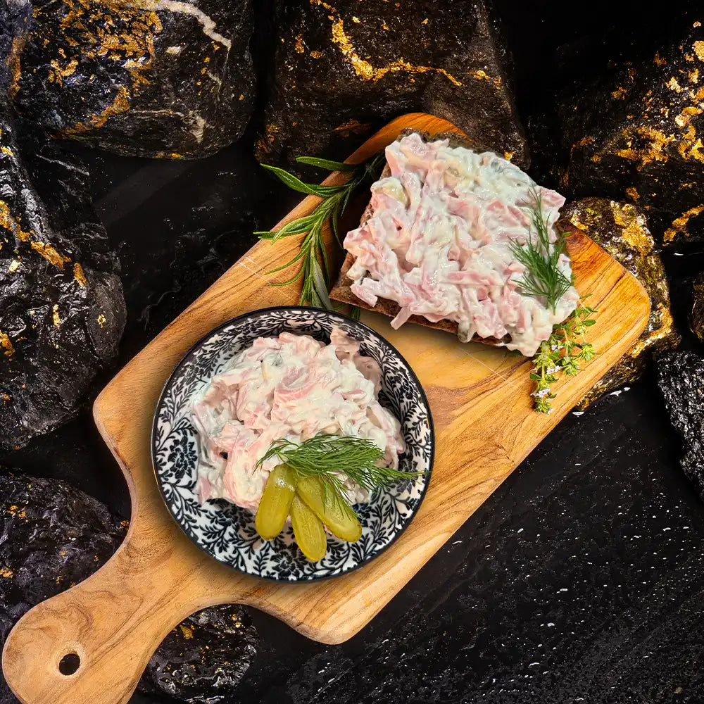Fleischsalat auf dunklem Teller neben Fleischsalat auf Brotscheibe auf einem Holzbrett. Dunkler Hintegrund mit Steinbrocken.