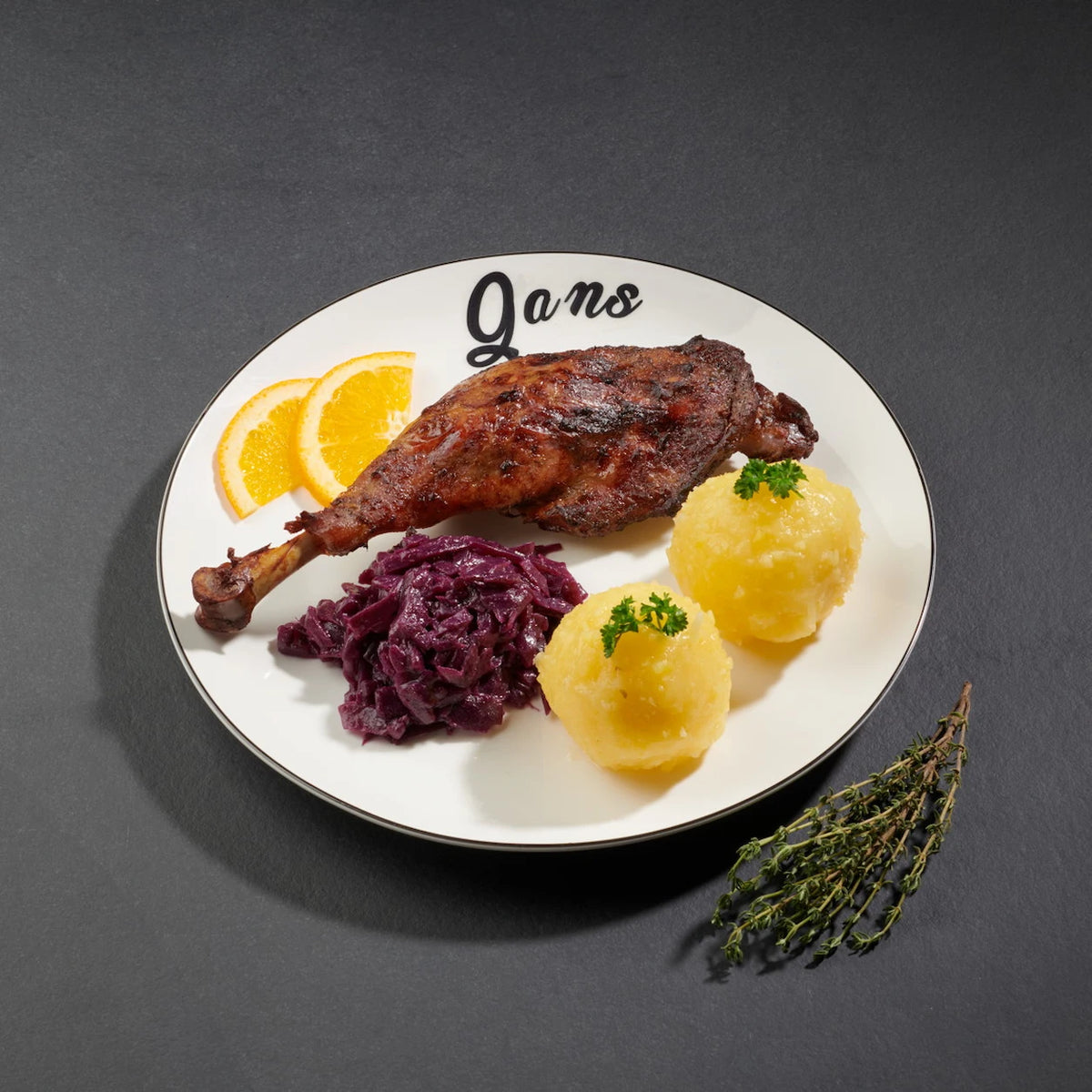 1 gegarte Gänsekeule auf einem weißen Teller mit Rotkohl und zwei Klößen und zwei Orangenscheiben.