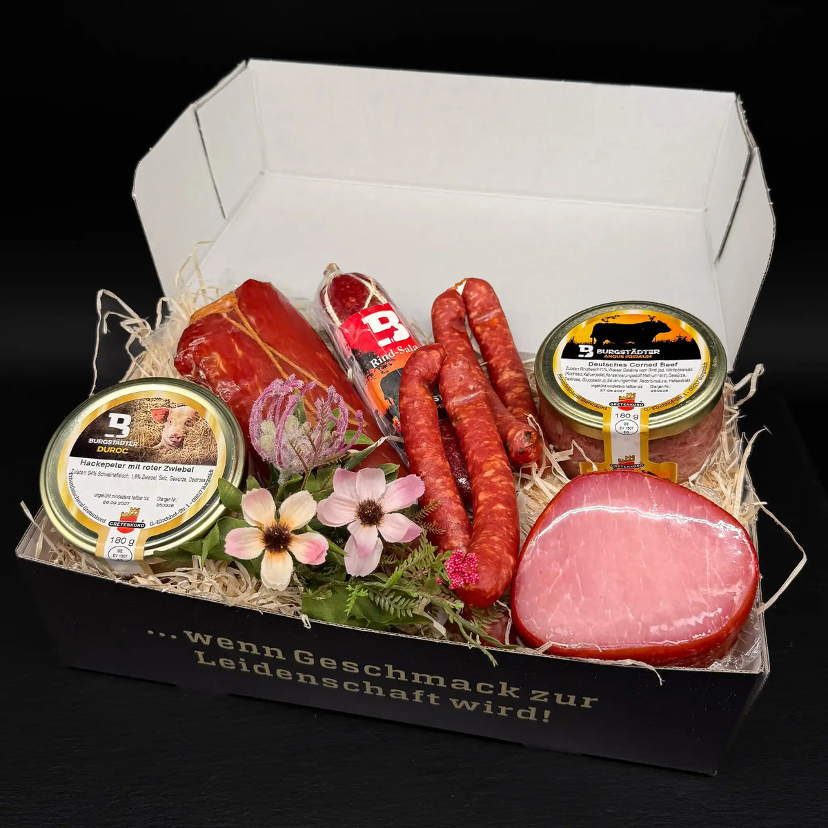 Eine rechteckige Box gefüllt mit einem Glas Corned Beef und einem Glas Hackepeter, 2 Paar Pfefferbeißern, einer Salami, einem geräucherten Schweinefilet und einem Lachsschinken.