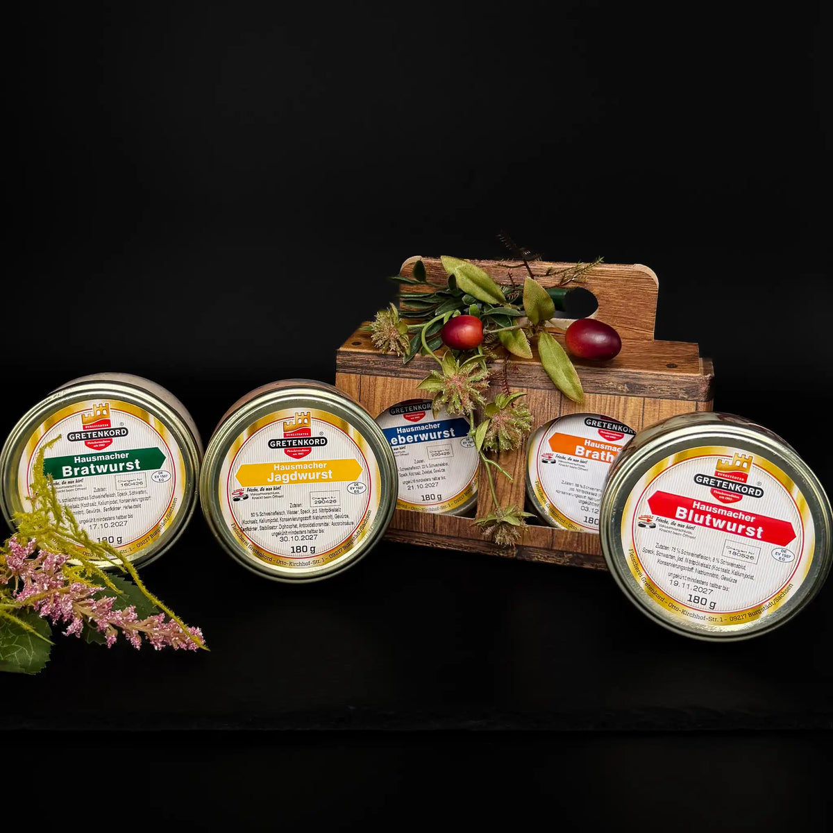 Eine rechteckige Holzbox gefüllt mit zwei Gläsern. Insgesamt sind zu sehen: Hausmacher Leberwurst, Bratwurst, Blutwurst, Brathaxe und Jagdwurst.