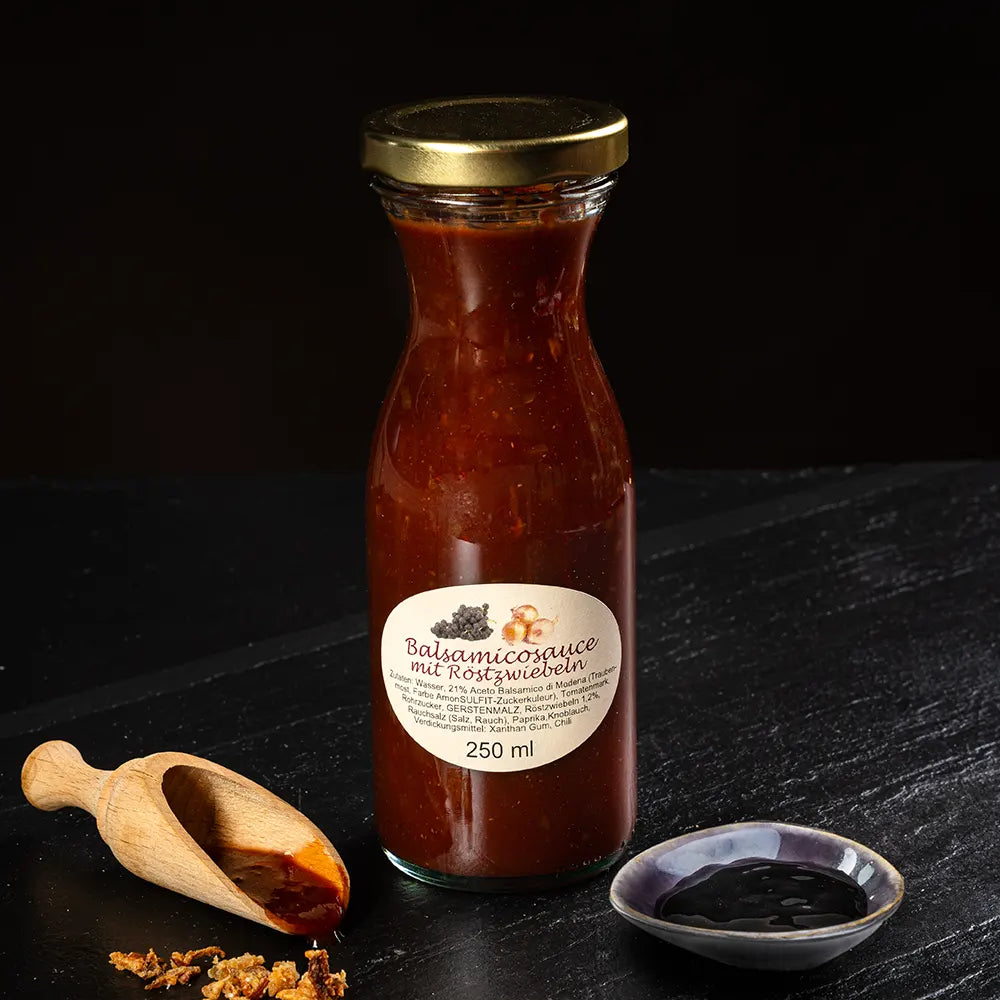 Rote Balsamicosauce mit Röstzwiebeln in kleiner Glasflasche neben kleinem Holzlöffel und Schälchen mit Sauce vor dunklem Hintergrund.
