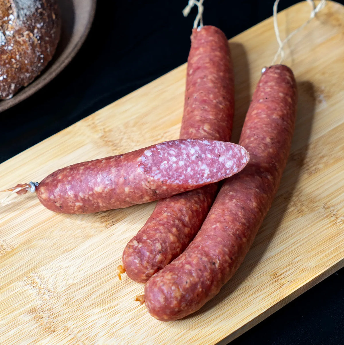 Eine schräg angeschnittene Salami und 2 ganze Salamis liegen quer zueinander auf einem hellem Holzbrett vor einem schwarzen Hintergrund.